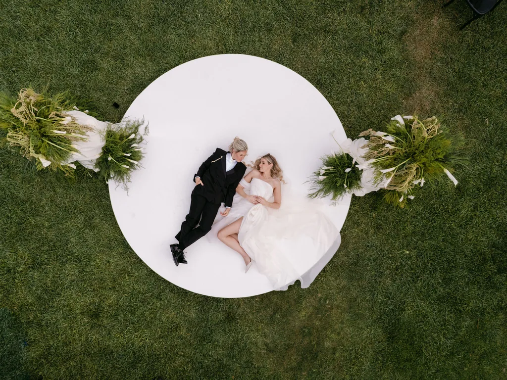 Emma and Whitney lying together on the white ceremony circle at Acre, seen from above, San José del Cabo
